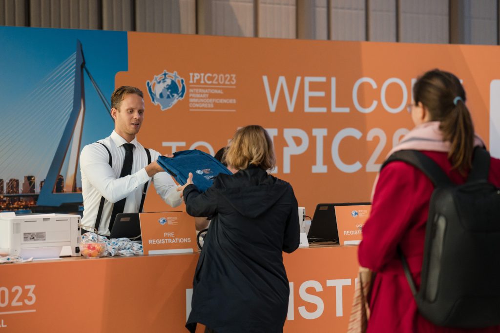 IPIC2025: A registration desk at an event, with a staff member handing a t-shirt to a visitor, set against a vibrant welcome banner.