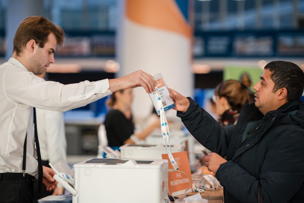 IPIC2025: At entrance desk, a staff member hands a ticket to a attendee in a winter coat, amidst a busy event atmosphere.